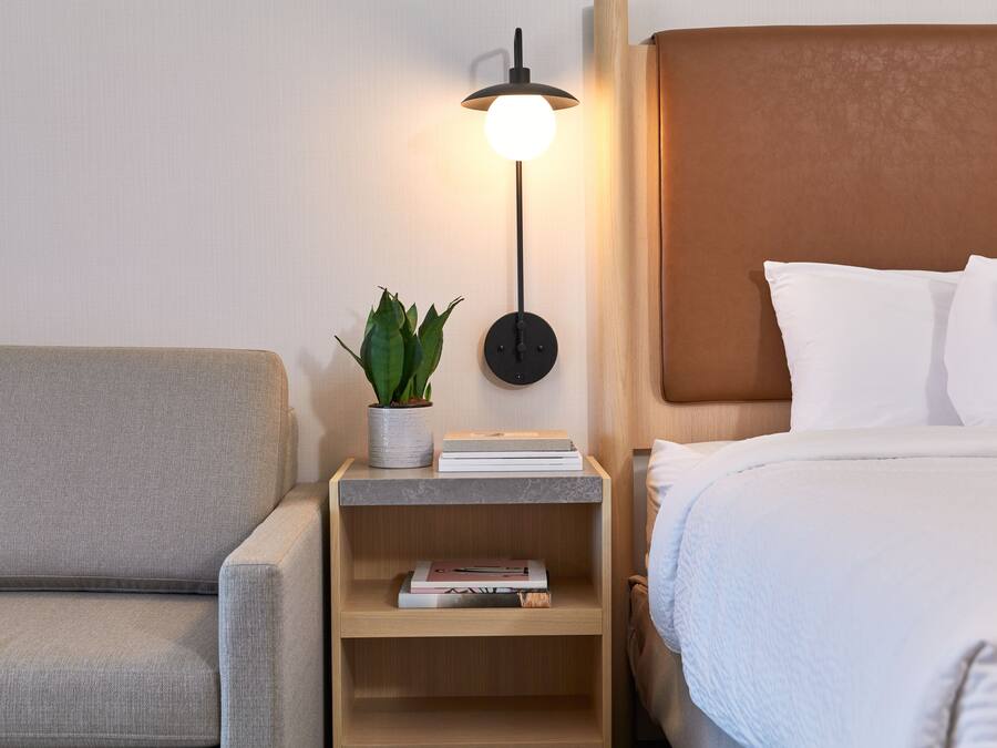 A neatly arranged hotel room with a bed, a bedside table with a lamp, a small plant, and a few books.