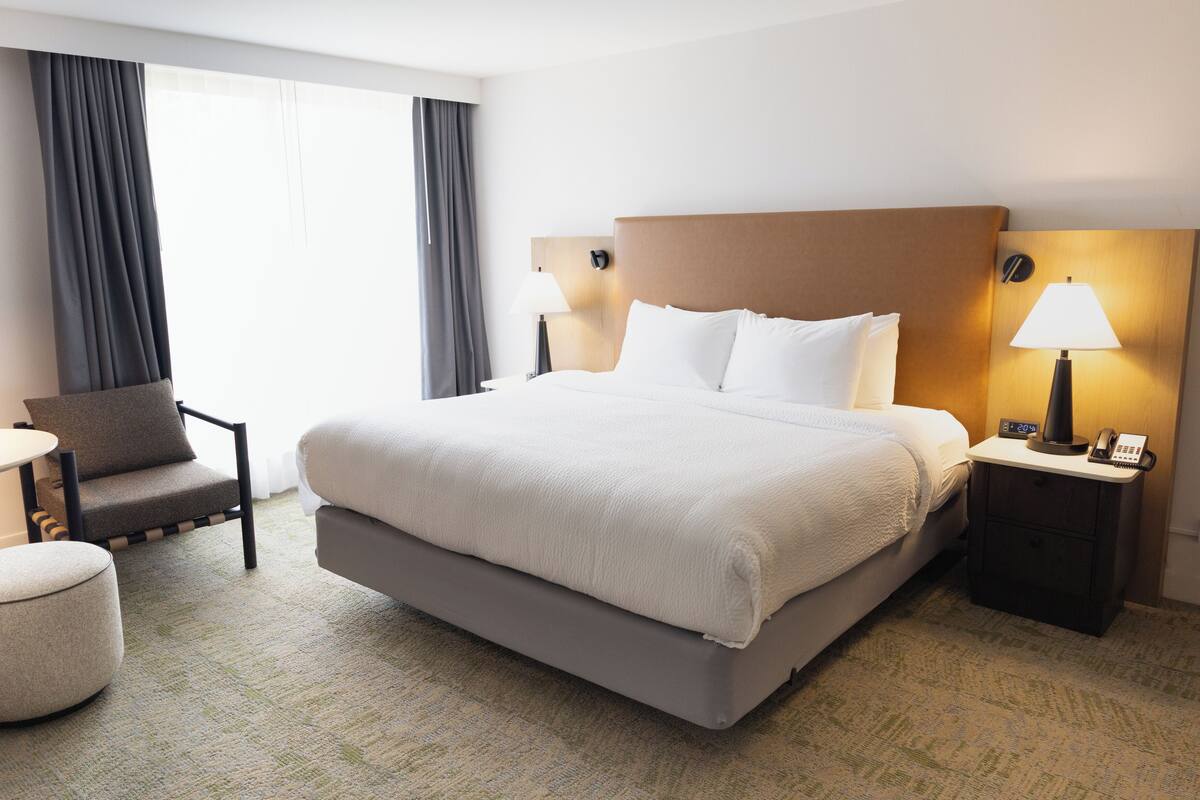 A hotel room with a large bed, a bedside table with a lamp, a chair, and a small table.