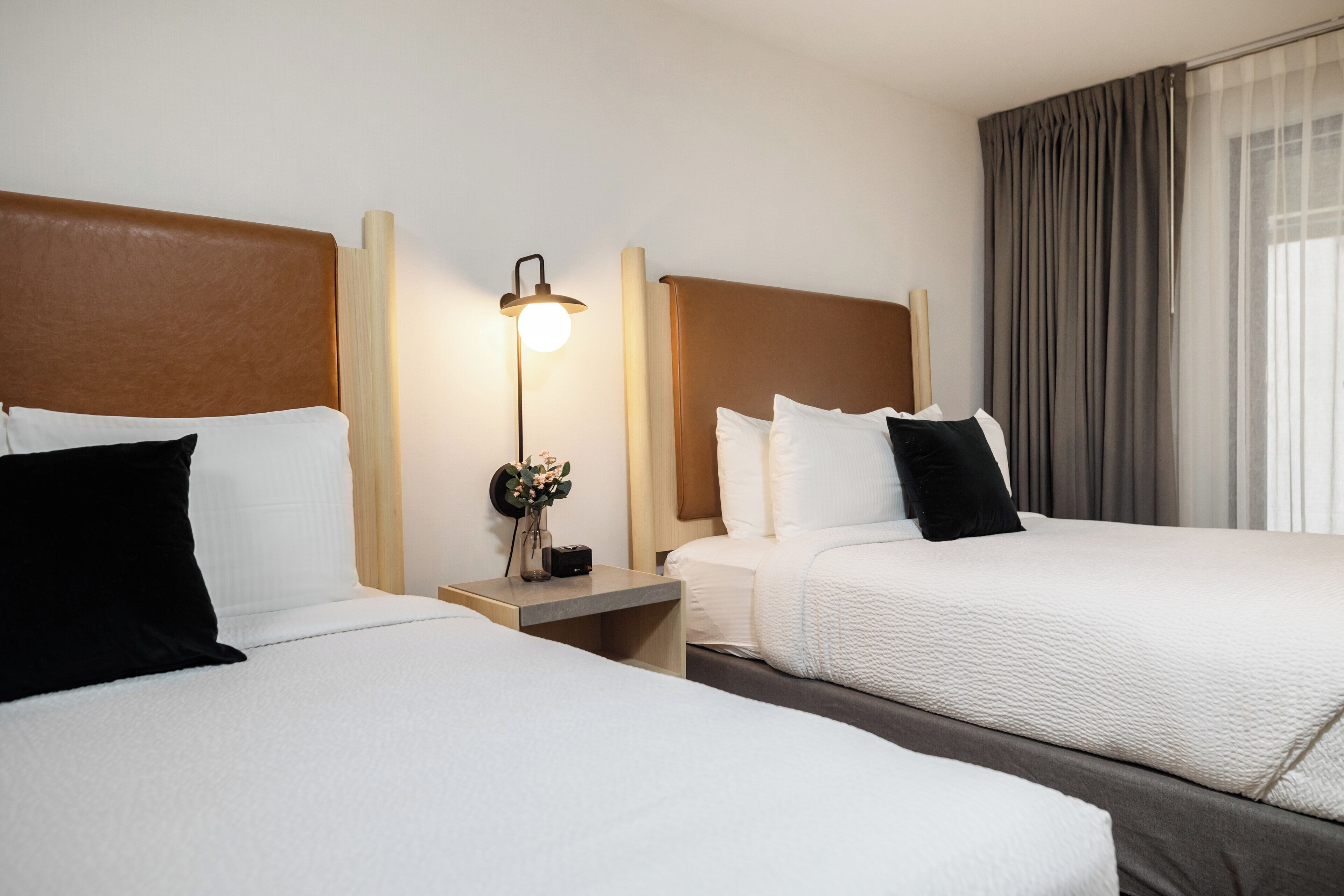 A hotel room with two beds, a nightstand with a lamp, and a large window with curtains.