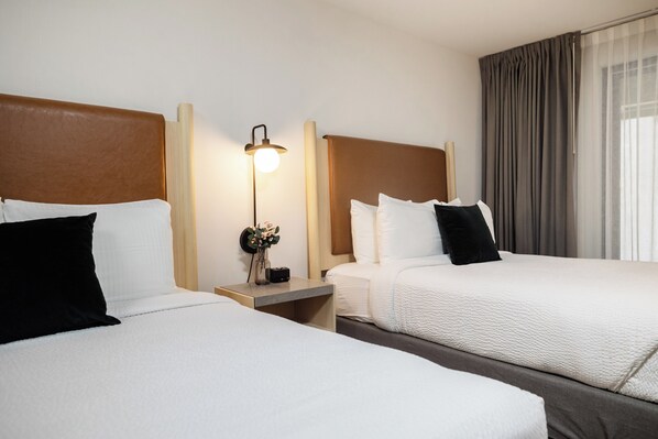 Habitación de hotel con dos camas, una mesita de noche con lámpara y un ventanal con cortinas.
