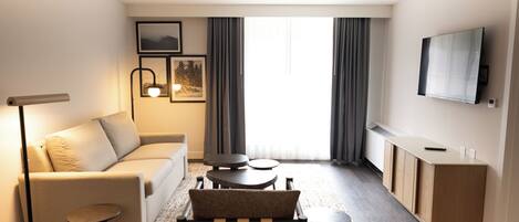 A modern hotel room with a sofa, a small round table, a chair, a desk, and a flat-screen TV on the wall.