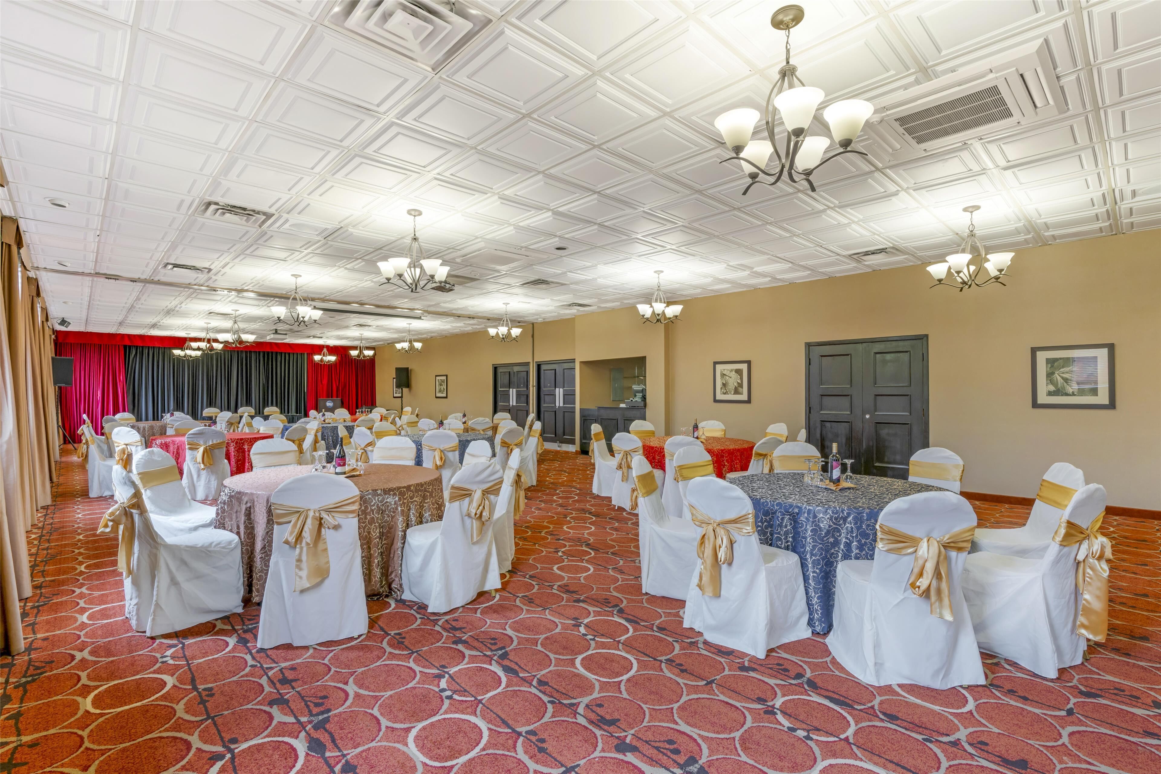 Un salón de banquetes con mesas redondas, sillas blancas con cintas doradas y un piso cubierto de alfombra roja.