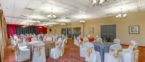 Un salón de banquetes con mesas redondas, sillas blancas con cintas doradas y un piso cubierto de alfombra roja.