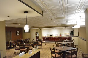 A hotel dining area with multiple tables and chairs, a kitchen in the background, and a ceiling with recessed lighting.