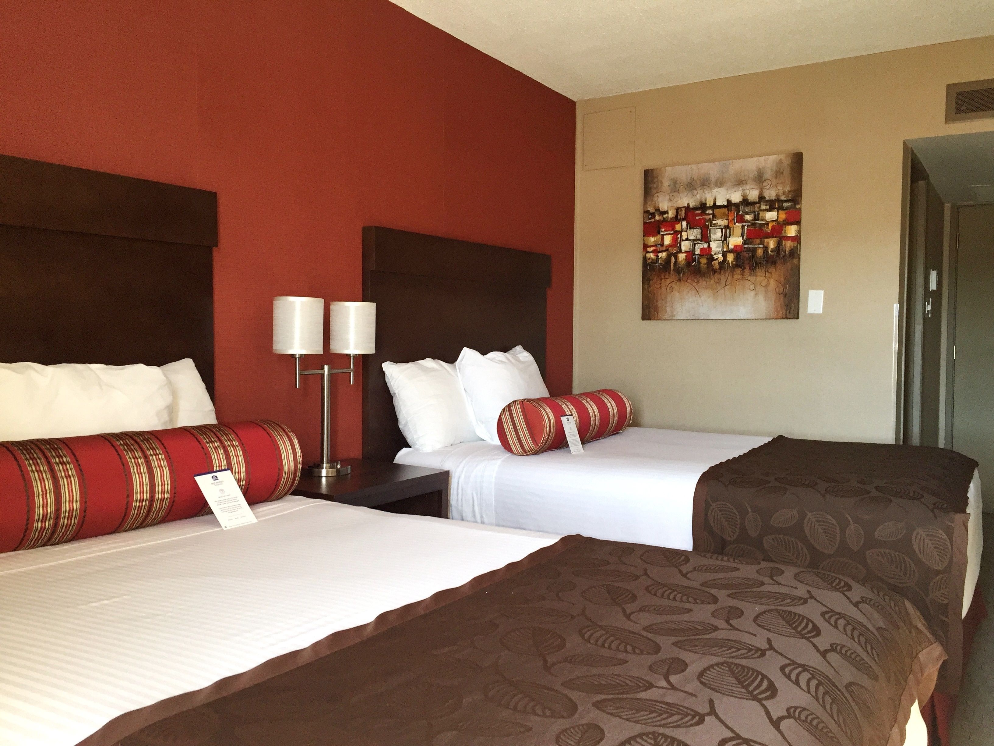 A hotel room with two beds, a red accent wall, a painting, and a nightstand with lamps.