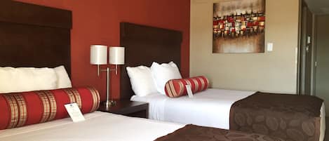 A hotel room with two beds, a red accent wall, a painting, and a nightstand with lamps.