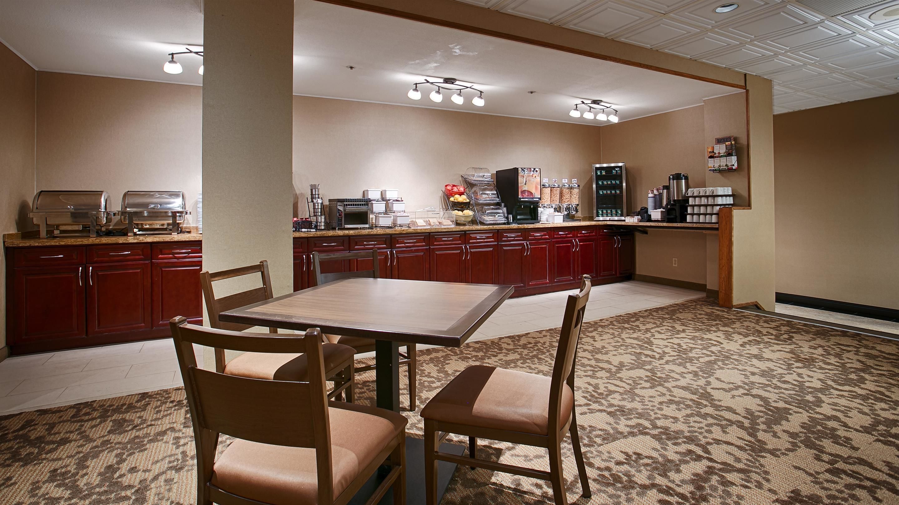 Habitación de hotel con zona de comedor que incluye una mesa y sillas de madera, kitchenette con cafetera y piso alfombrado.