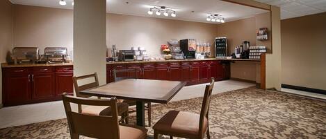Habitación de hotel con zona de comedor que incluye una mesa y sillas de madera, kitchenette con cafetera y piso alfombrado.