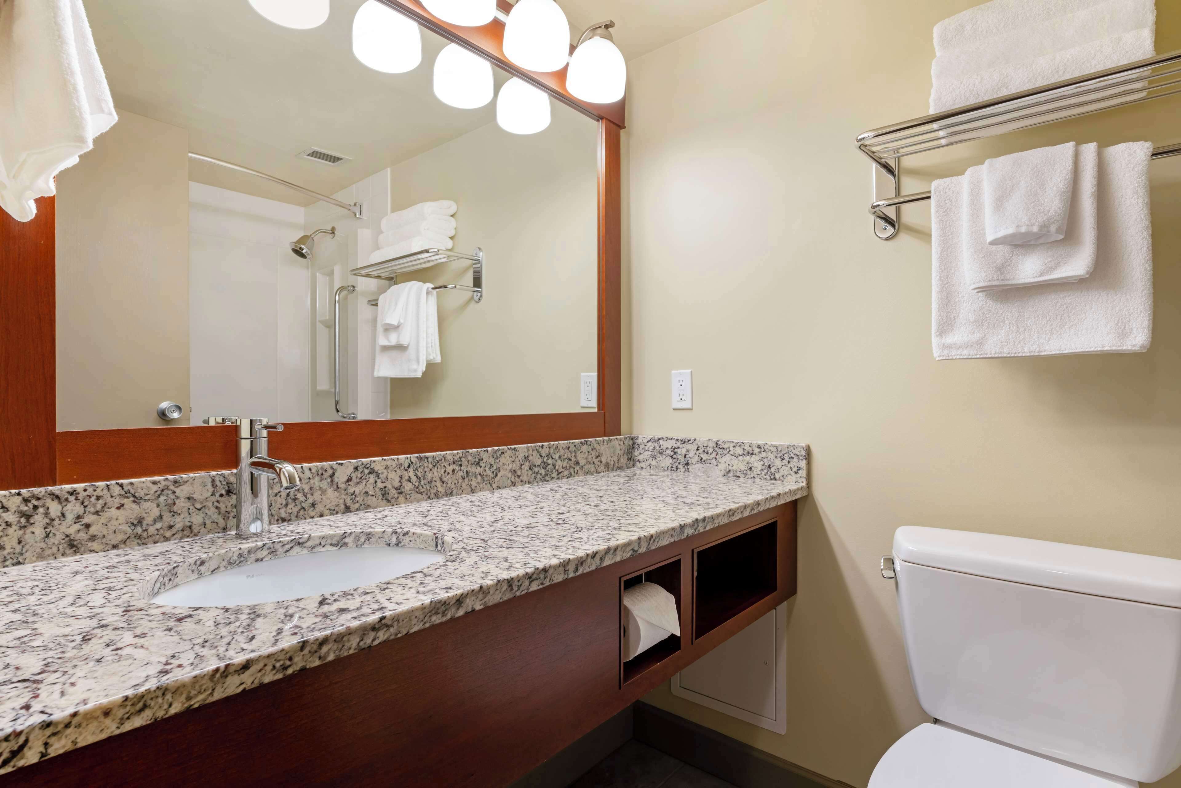 A bathroom with a granite countertop sink, a mirror with lights, a towel rack, and a toilet.