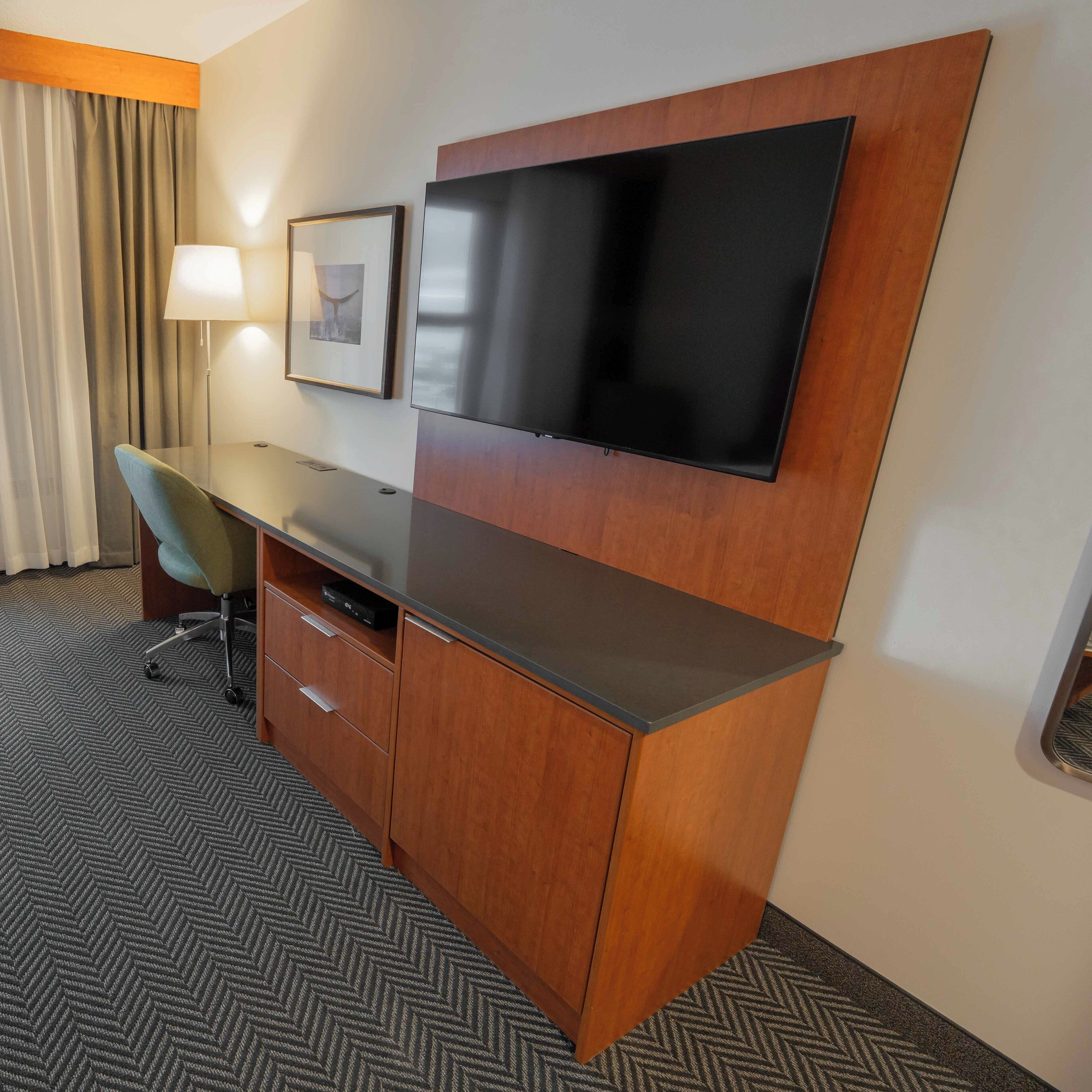 A hotel room with a wooden desk, a flat-screen TV mounted on the wall, a chair, a lamp, and a framed picture.