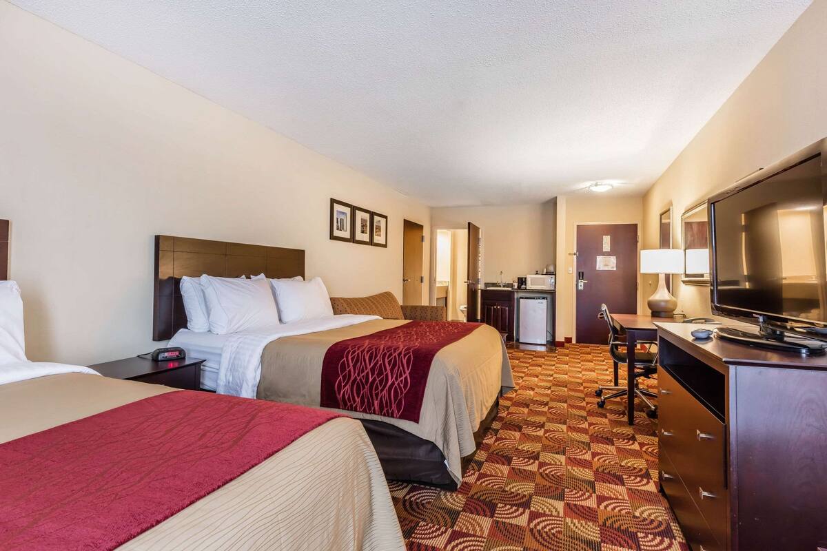 A hotel room with two beds, a desk with a TV, a small kitchenette, and a carpeted floor.