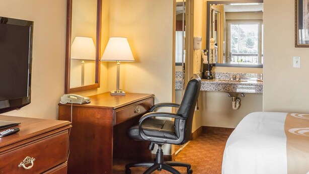 A hotel room with a wooden desk, two lamps, a black office chair, a bed, a large mirror, a bathroom with a sink and vanity, and a television. - Quality Inn Calera I-65 exit 231 (Calera)