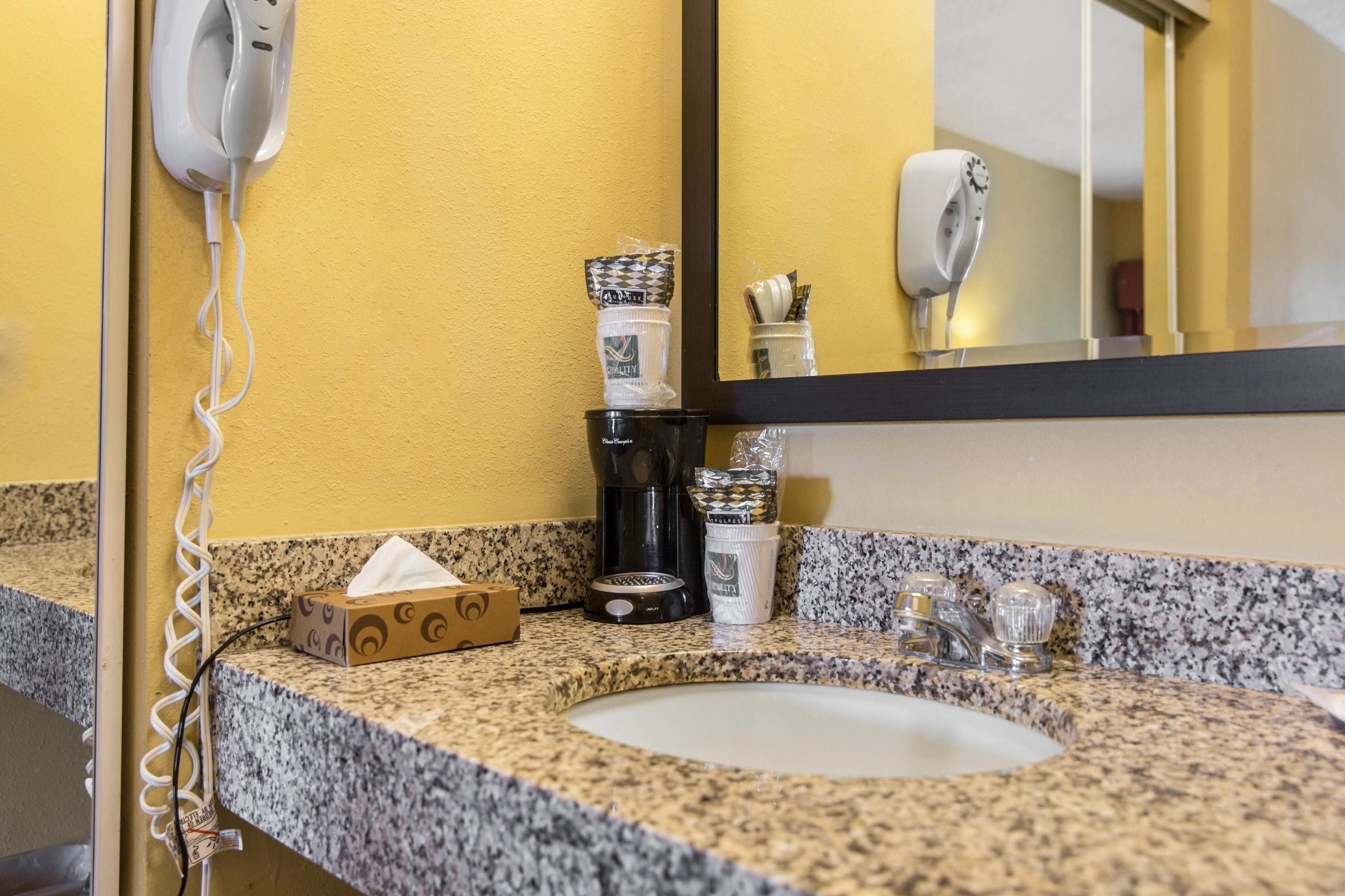 A bathroom with a granite countertop, a sink, a mirror, a hairdryer, a coffee maker, and toiletries.