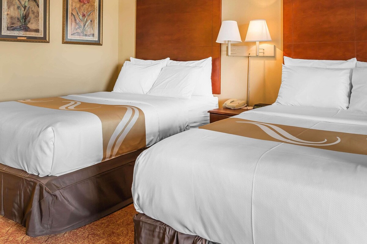 A hotel room with two beds, each with white bedding and brown headboards. There are two wall-mounted lamps and a telephone on the nightstand.