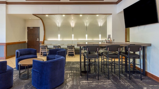Un hall d’hôtel avec des sièges bleus, un coin bar et un grand écran de télévision.