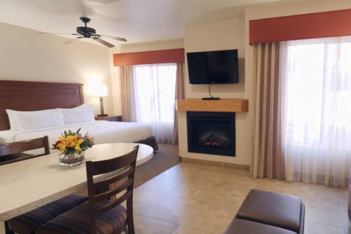 A hotel room with a bed, a TV mounted on the wall, a fireplace, a dining area with a table and chairs, and a sofa.