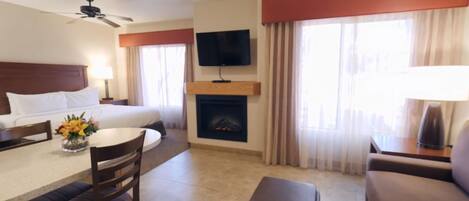 A hotel room with a bed, a TV mounted on the wall, a fireplace, a dining area with a table and chairs, and a sofa.