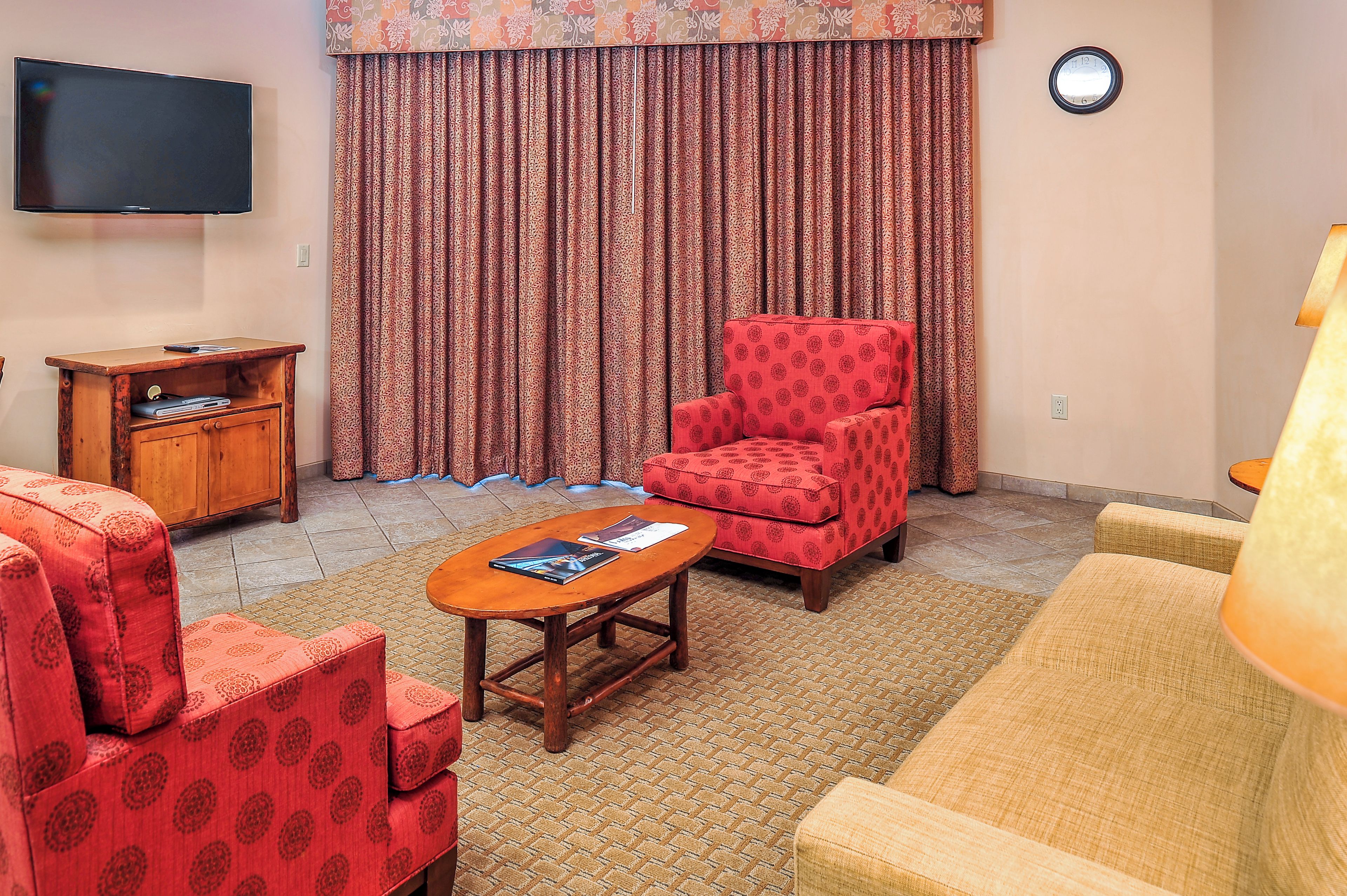 A living room with a flat-screen TV, a red patterned armchair, a wooden cabinet, and a patterned rug.