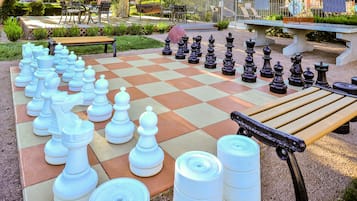 Un tablero de ajedrez grande al aire libre en un jardín, con mesas y sillas al fondo.