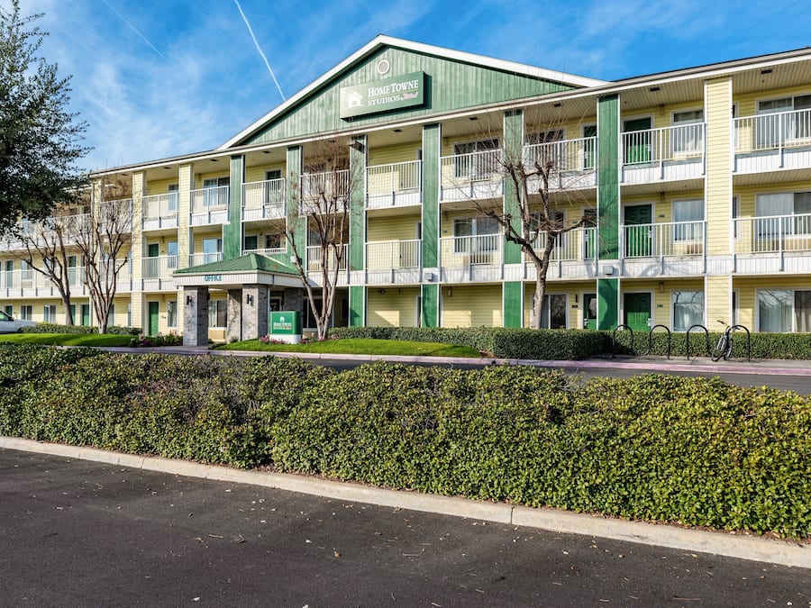 A multi-story hotel with a green and yellow facade, multiple balconies, and a parking area in front.