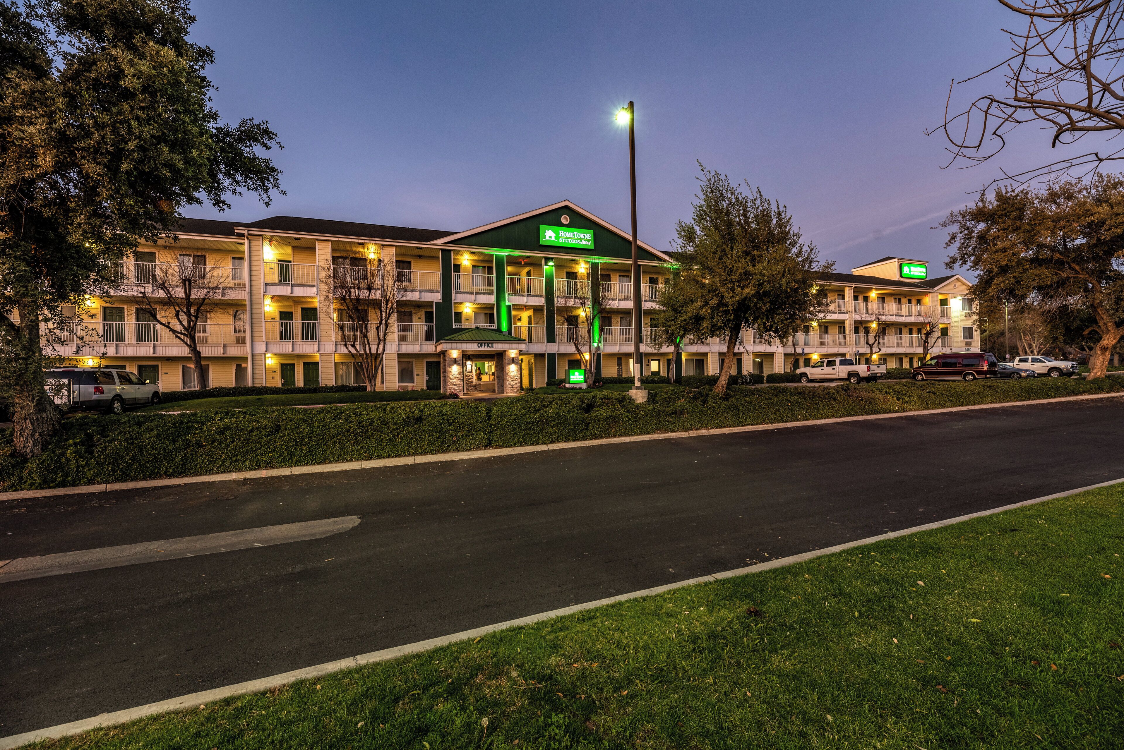 Ein mehrstöckiges Hotel mit einem grünen Schild, auf dem 'Home Town Studios' steht, und einem Parkplatz davor.