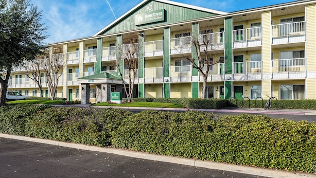 A multi-story hotel with a green and yellow facade, multiple balconies, and a well-maintained hedge in front. - HomeTowne Studios by Red Roof Fresno - West (Fresno)