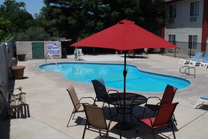 A swimming pool with lounge chairs and a table, surrounded by a concrete deck and a building in the background.