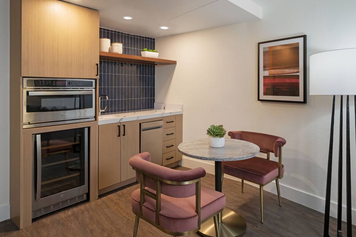 A compact kitchen with built-in appliances, a dining area with a round table and two chairs, and a wall-mounted lamp.