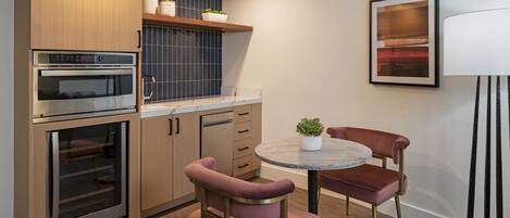 A compact kitchen with built-in appliances, a dining area with a round table and two chairs, and a wall-mounted lamp.