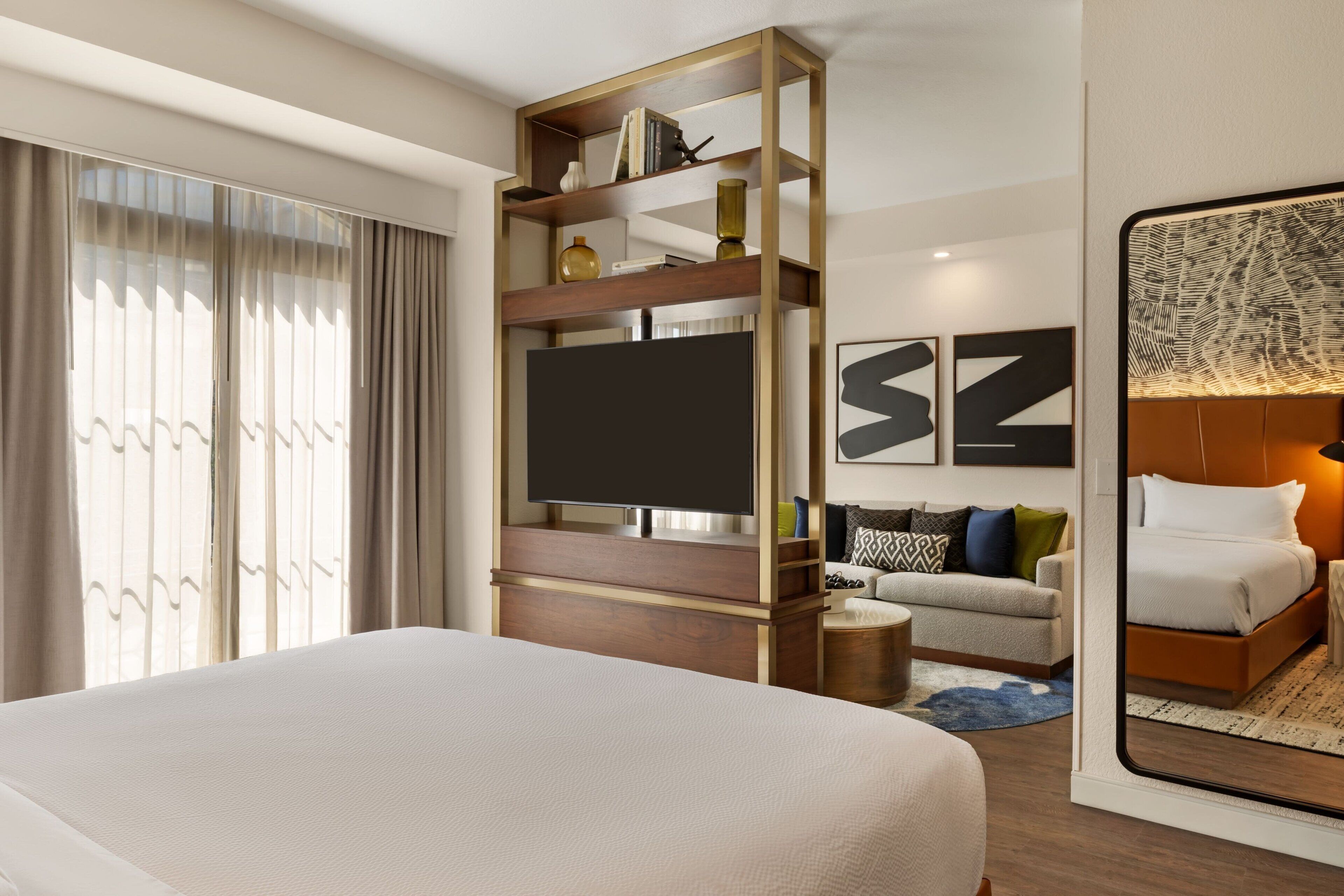 Une chambre d’hôtel moderne avec un grand lit, un téléviseur posé sur une étagère en bois, un canapé et un grand miroir qui reflète le lit.