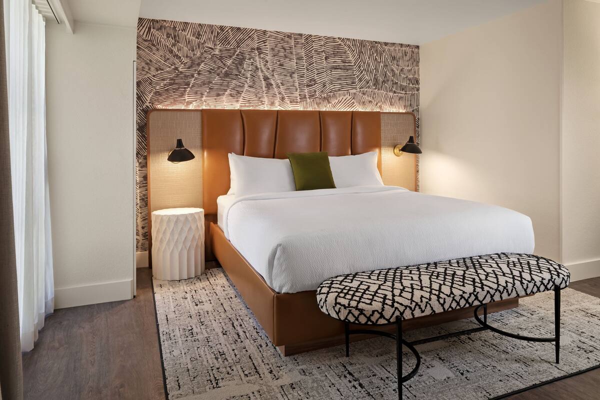 A modern bedroom with a large bed, a headboard with a leaf pattern, two bedside lamps, a round nightstand, and a patterned bench.