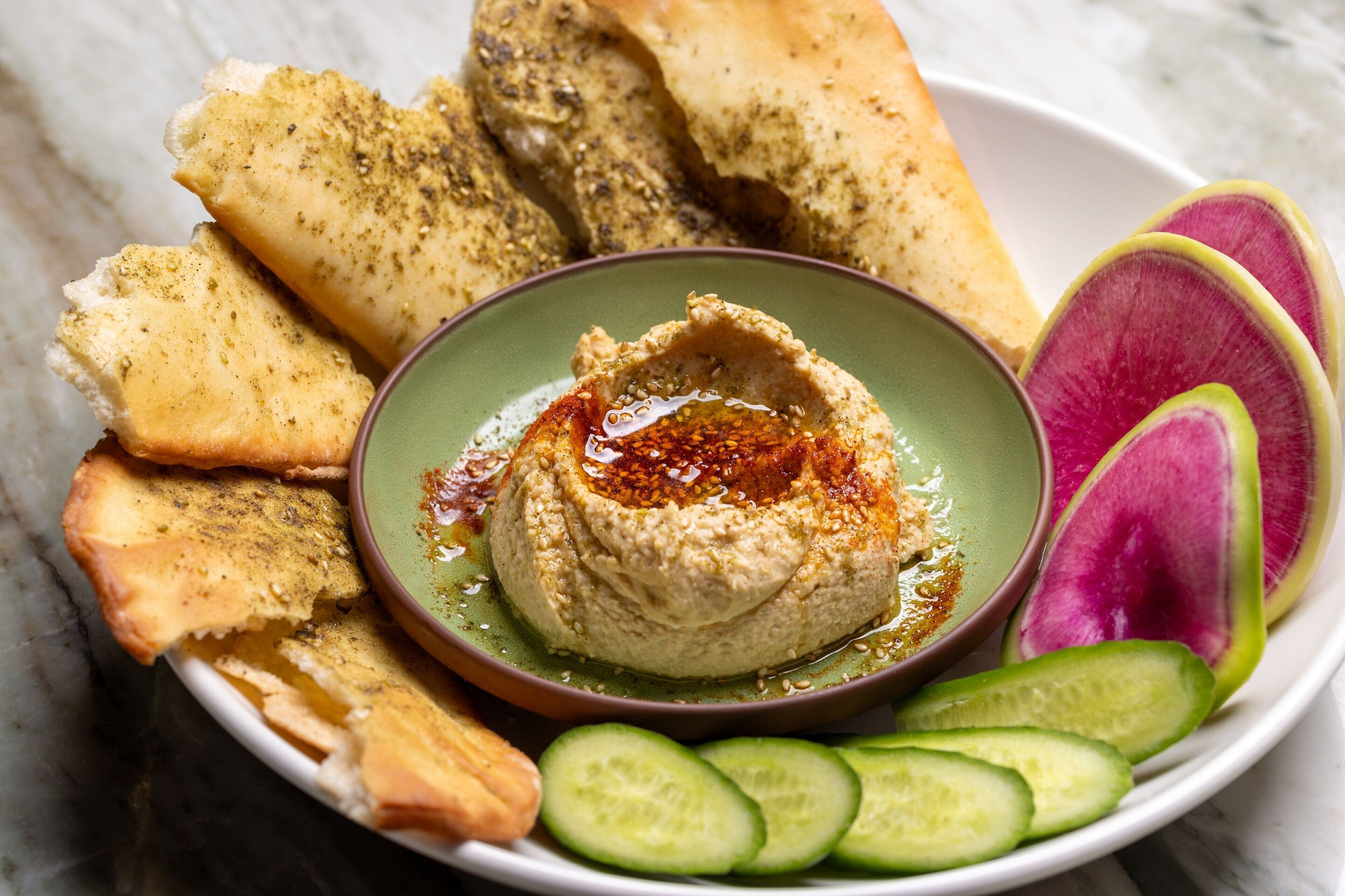 A plate of hummus with cucumber slices and pita bread.