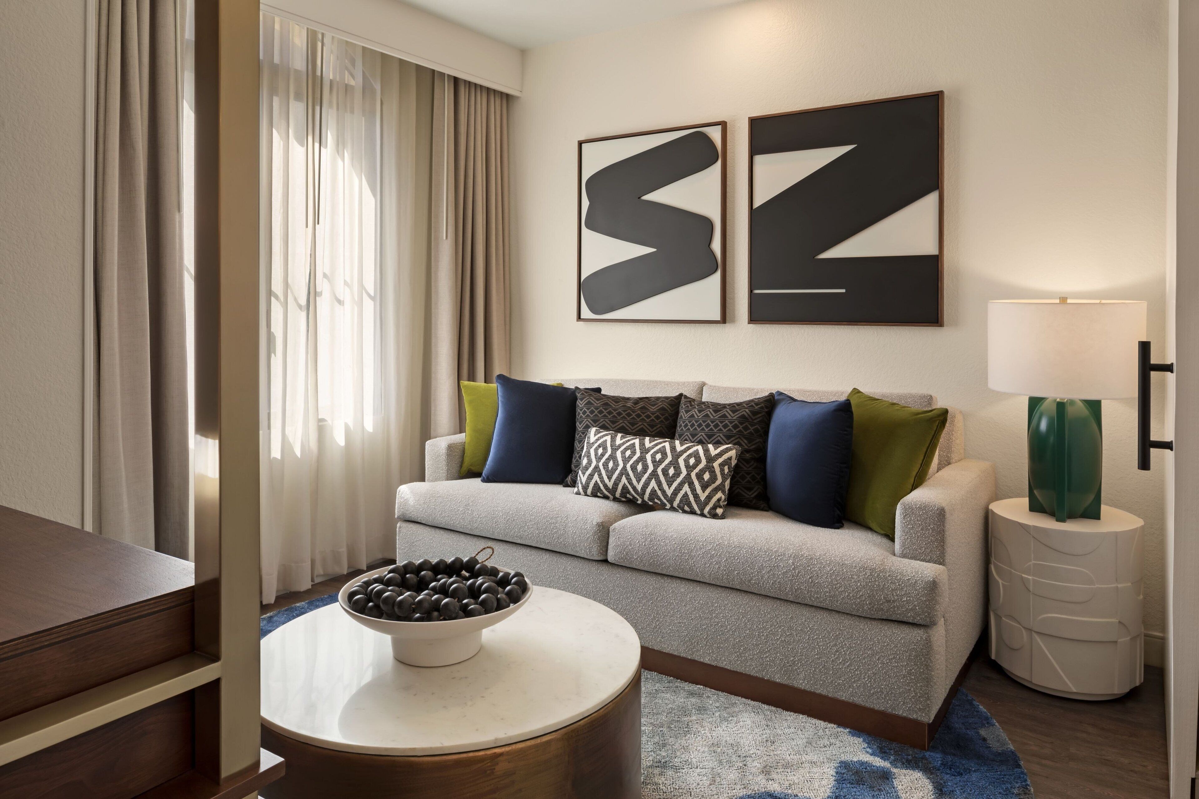 A modern living room with a grey sofa, patterned cushions, a round coffee table with a bowl of fruit, and abstract wall art.