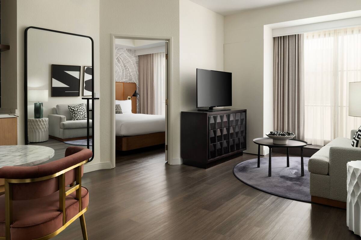 A modern hotel room with a bed, a TV, a seating area with a table, and a large window with curtains.