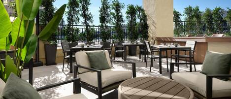 An outdoor patio area with multiple tables and chairs, surrounded by potted plants and trees.