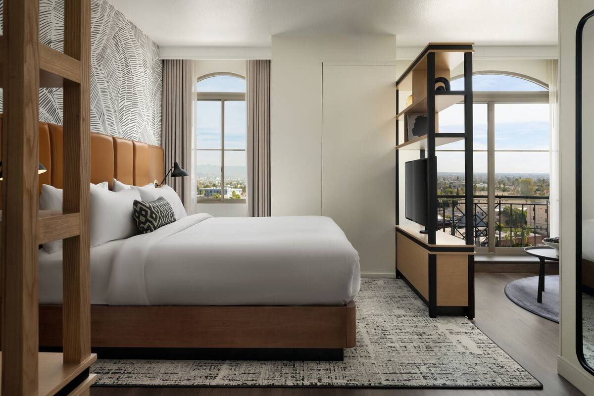 A modern hotel room with a large bed, a TV, a balcony with a view, and a wooden headboard.
