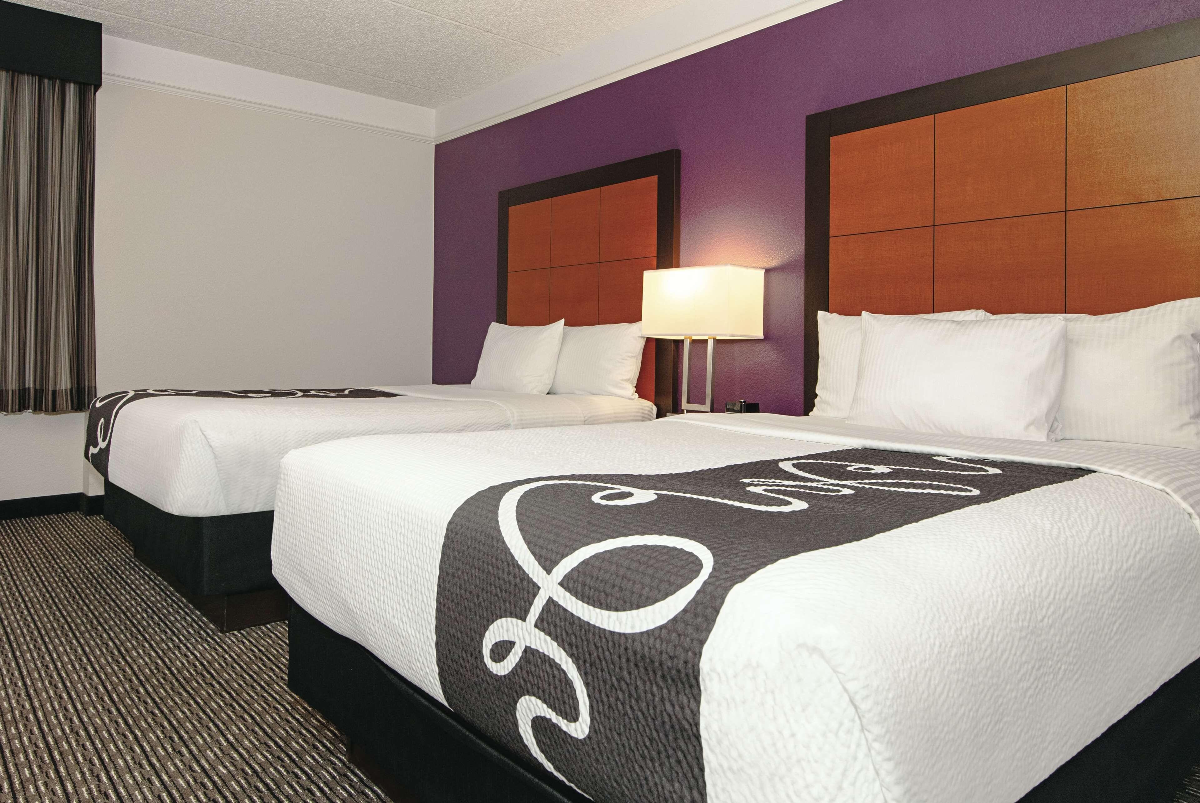 A hotel room with two beds, a headboard with a wooden panel design, a lamp on the nightstand, and a carpeted floor.