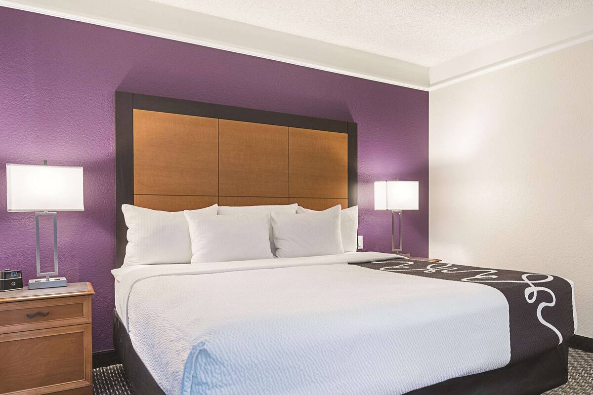 Hotellihuone, jossa on suuri sänky, kaksi yöpöytävalaisinta, puinen pääty ja violetit seinät.