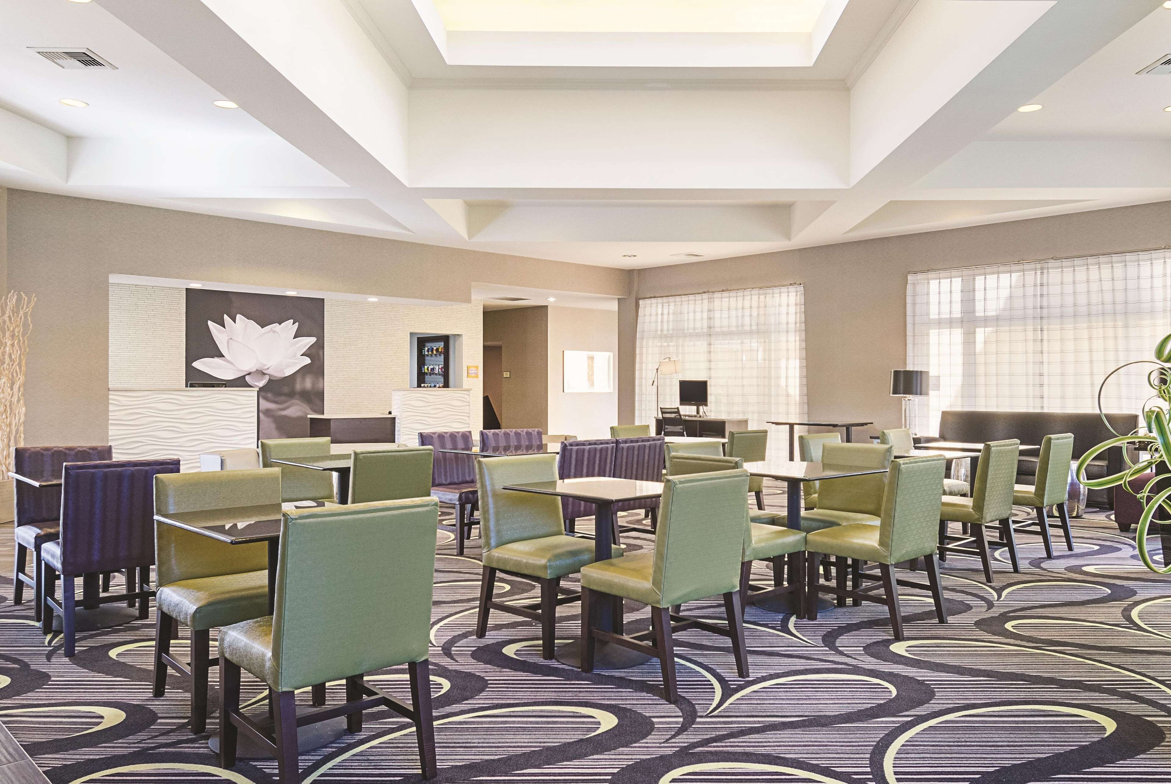 A hotel dining area with multiple tables and chairs, a large floral artwork on the wall, and a patterned carpet.