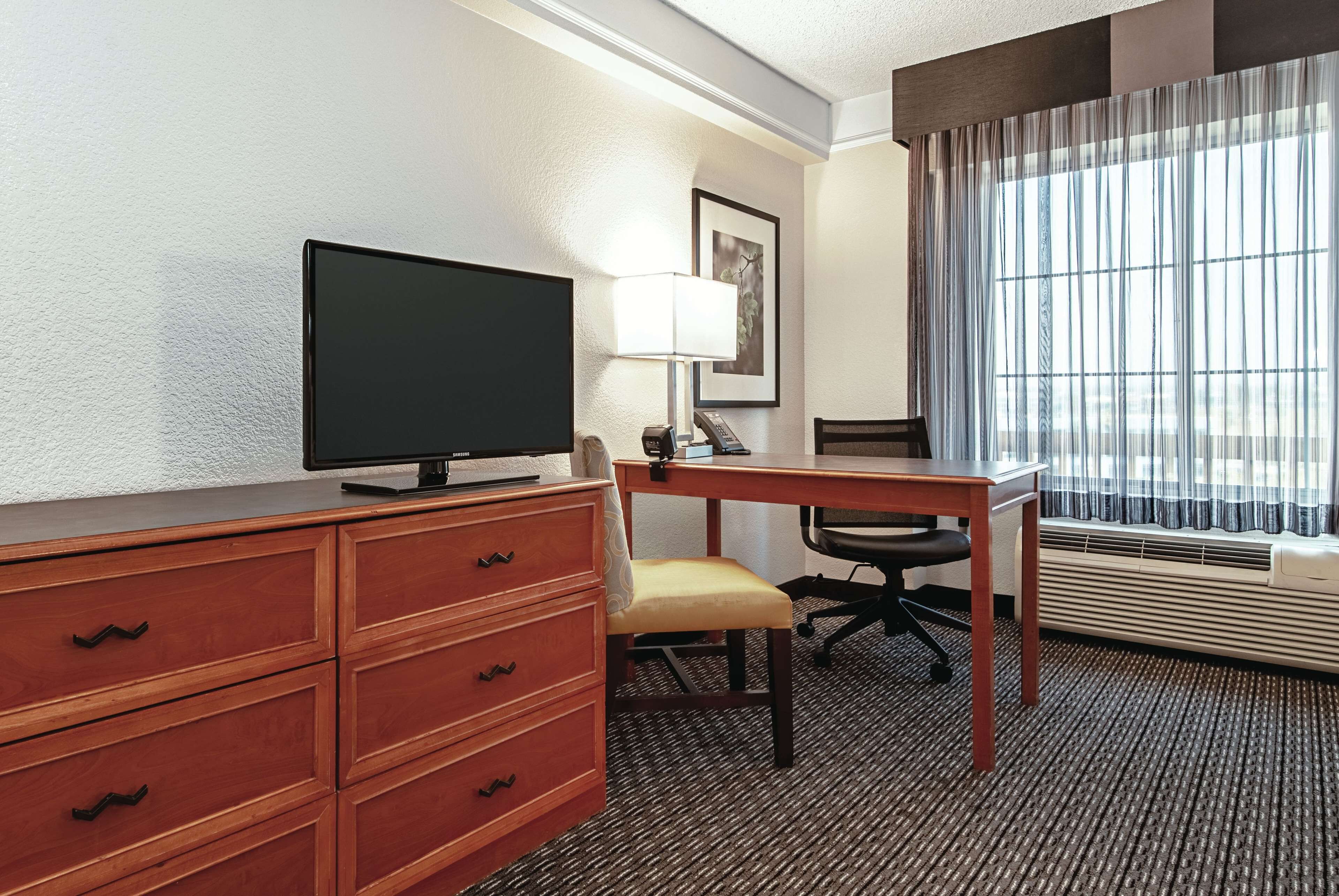 Habitación de hotel con escritorio, silla, televisor y un amplio ventanal con cortinas.