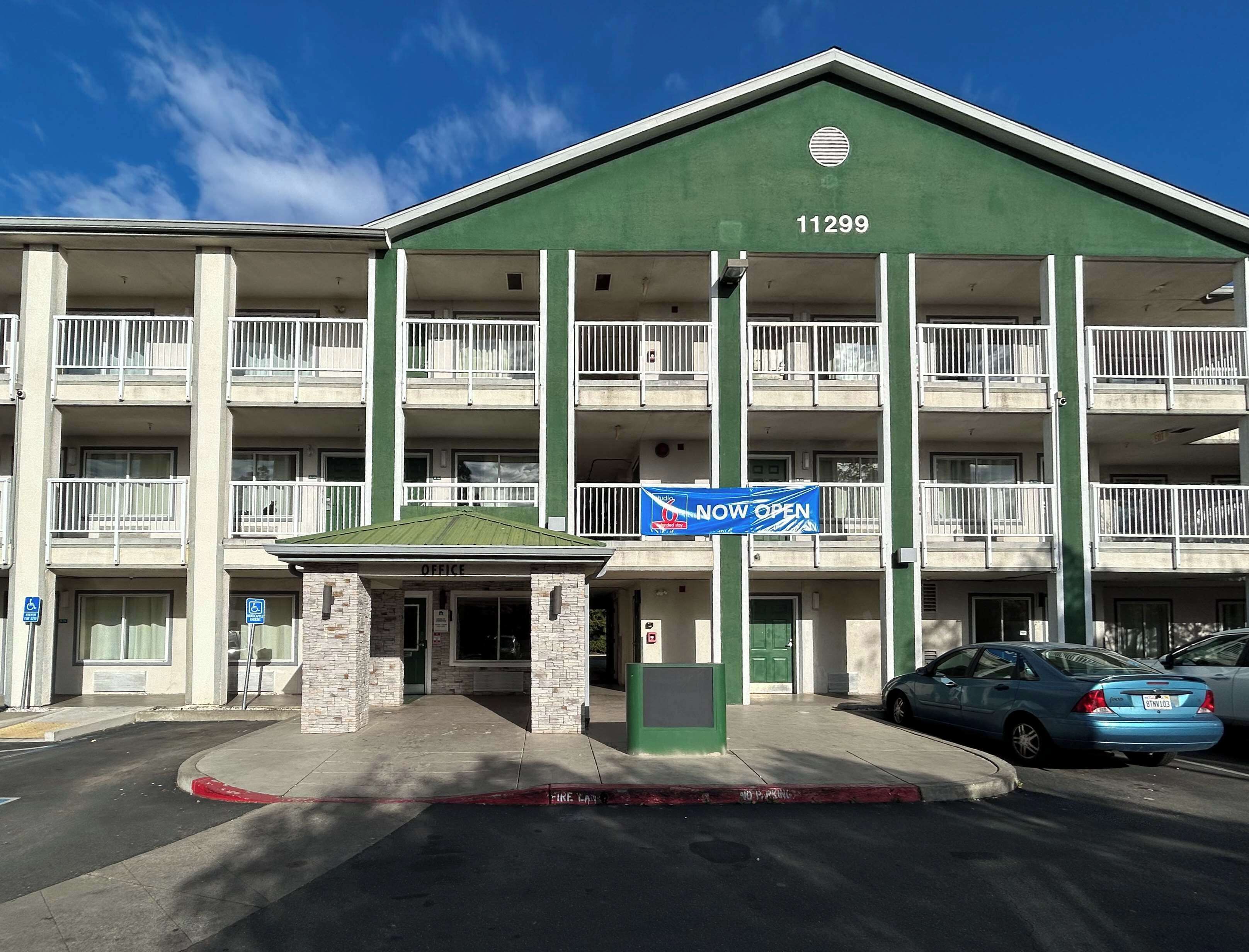 Ein mehrgeschossiges Hotel mit grüner Fassade, Nummer 11299, mit Balkonen, einem Eingangsbereich für Büros und einem blauen Schild mit der Aufschrift 'NOW OPEN'.