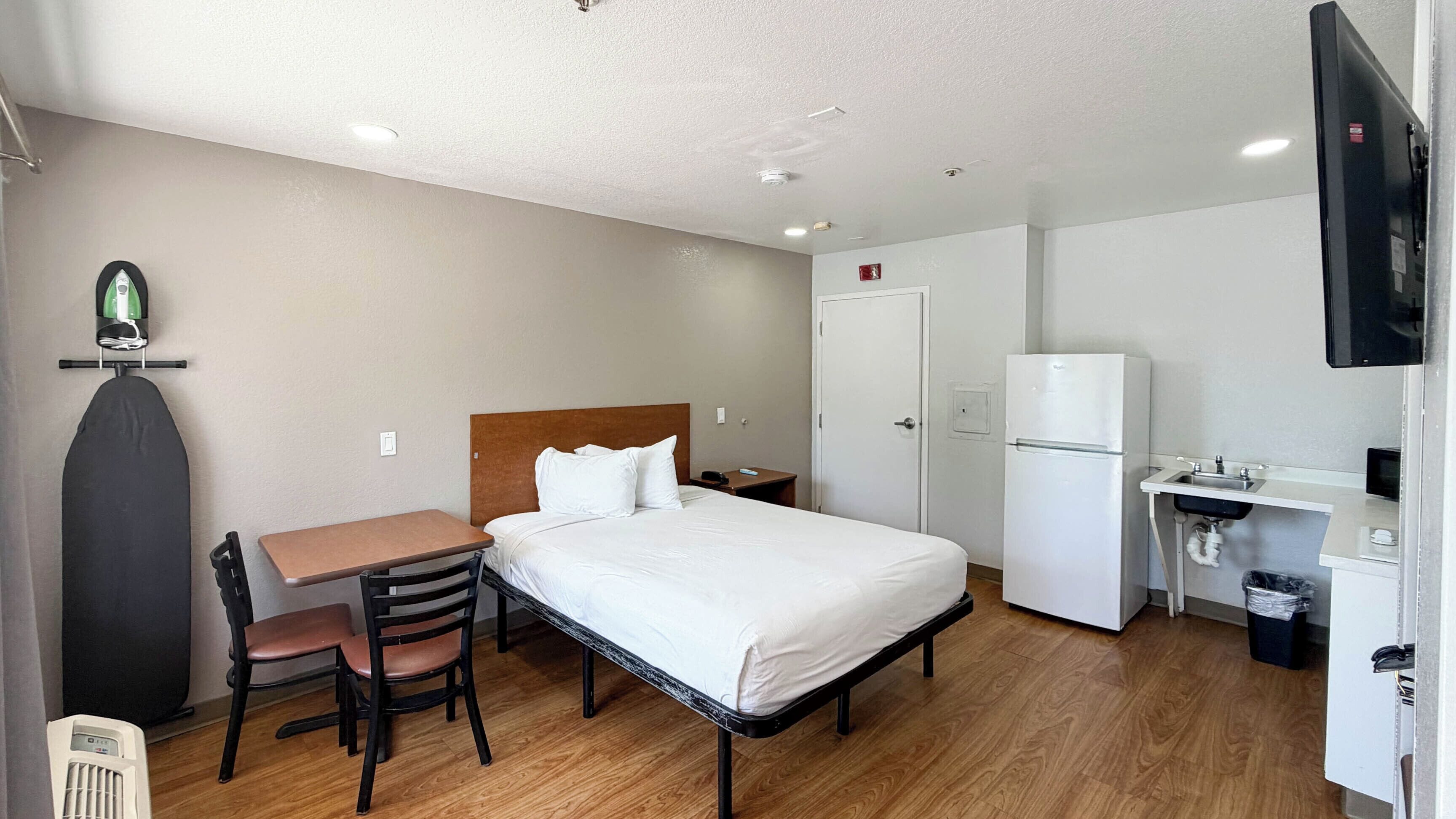 A hotel room with a bed, a desk, a chair, a small table, a refrigerator, a TV, and a trash bin.