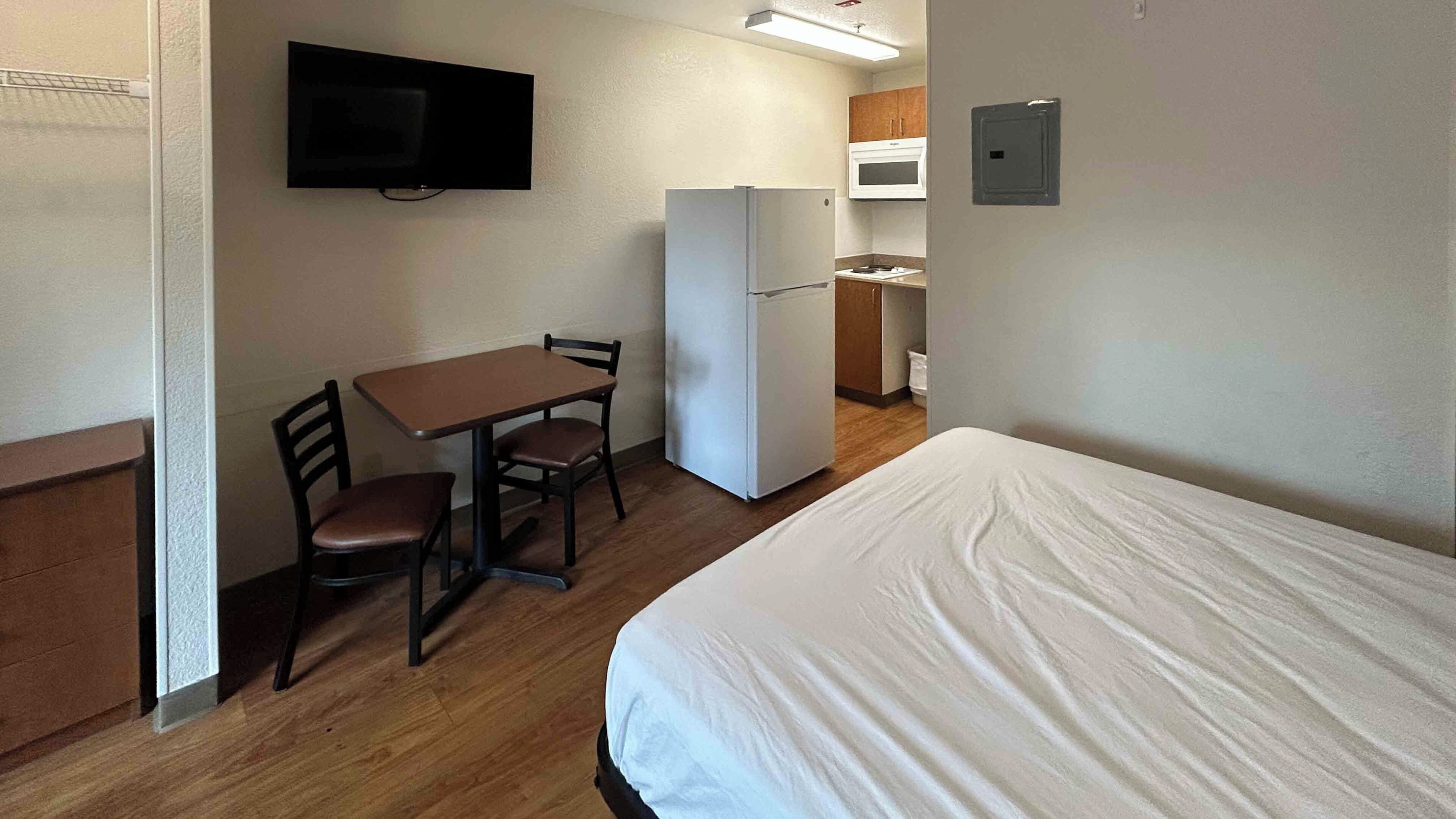 Una habitación pequeña con cama, una mesa con dos sillas, un refrigerador, un microondas y un televisor montado en la pared.
