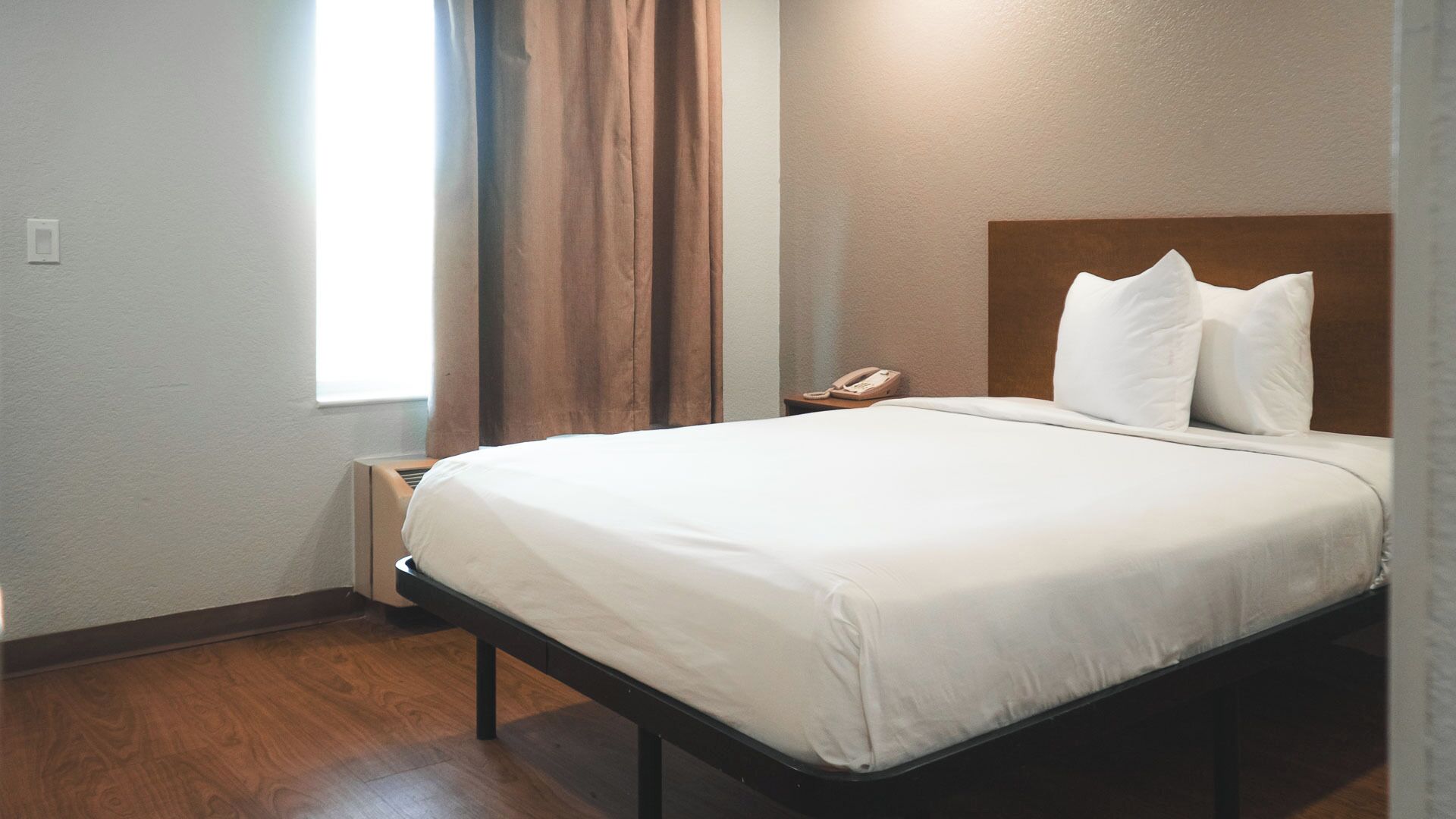 A neatly made bed in a hotel room with a wooden headboard, a bedside telephone, and a window with curtains.