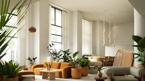 A spacious living room with a sofa, armchair, and coffee table, surrounded by potted plants and a large window. - Sandbourne Santa Monica, Autograph Collection (Santa Monica)