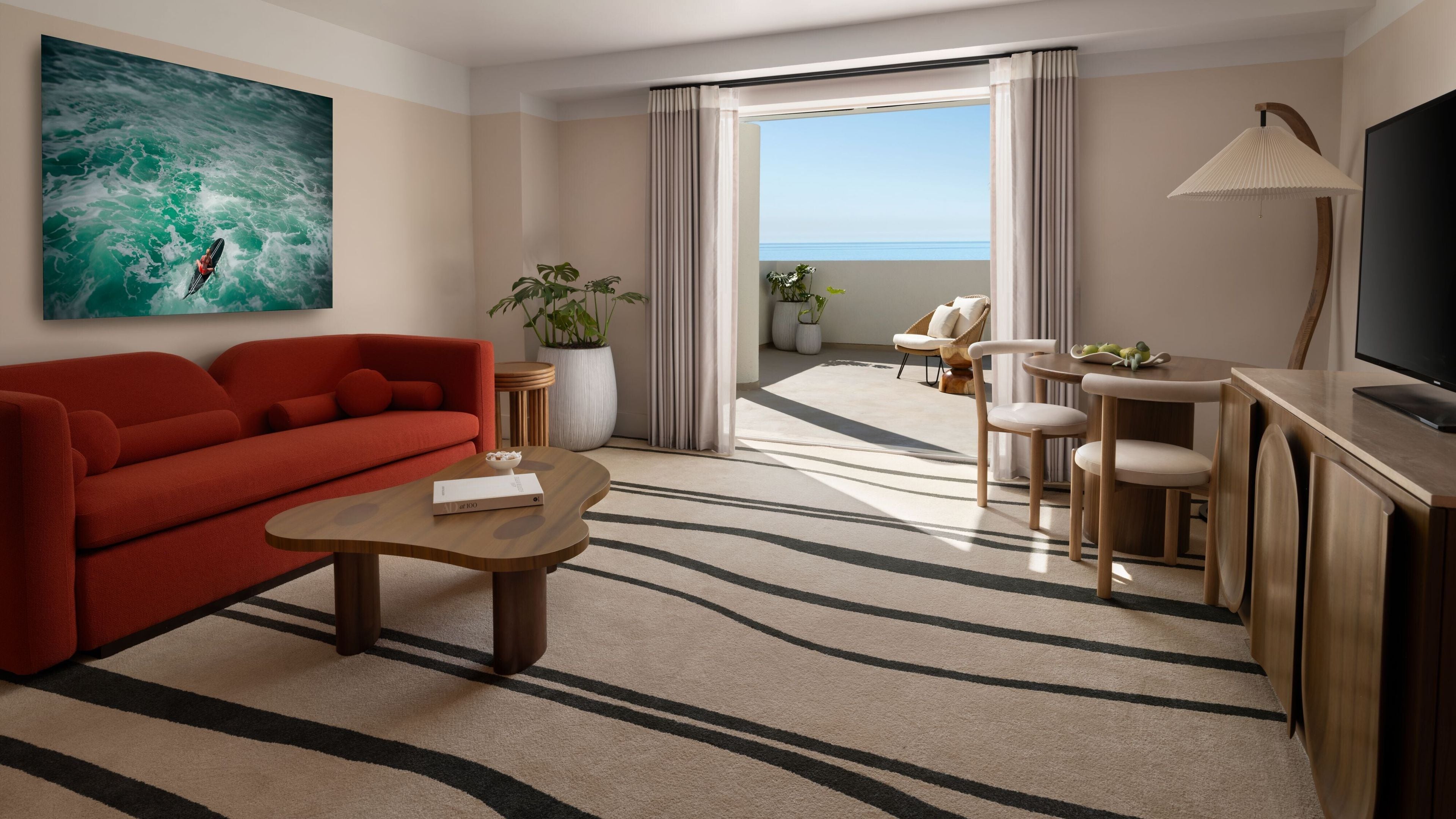 Une chambre d’hôtel avec un canapé rouge, une table basse en bois, un téléviseur, un coin repas avec une table et des chaises, une grande fenêtre donnant sur la mer et une plante en pot.
