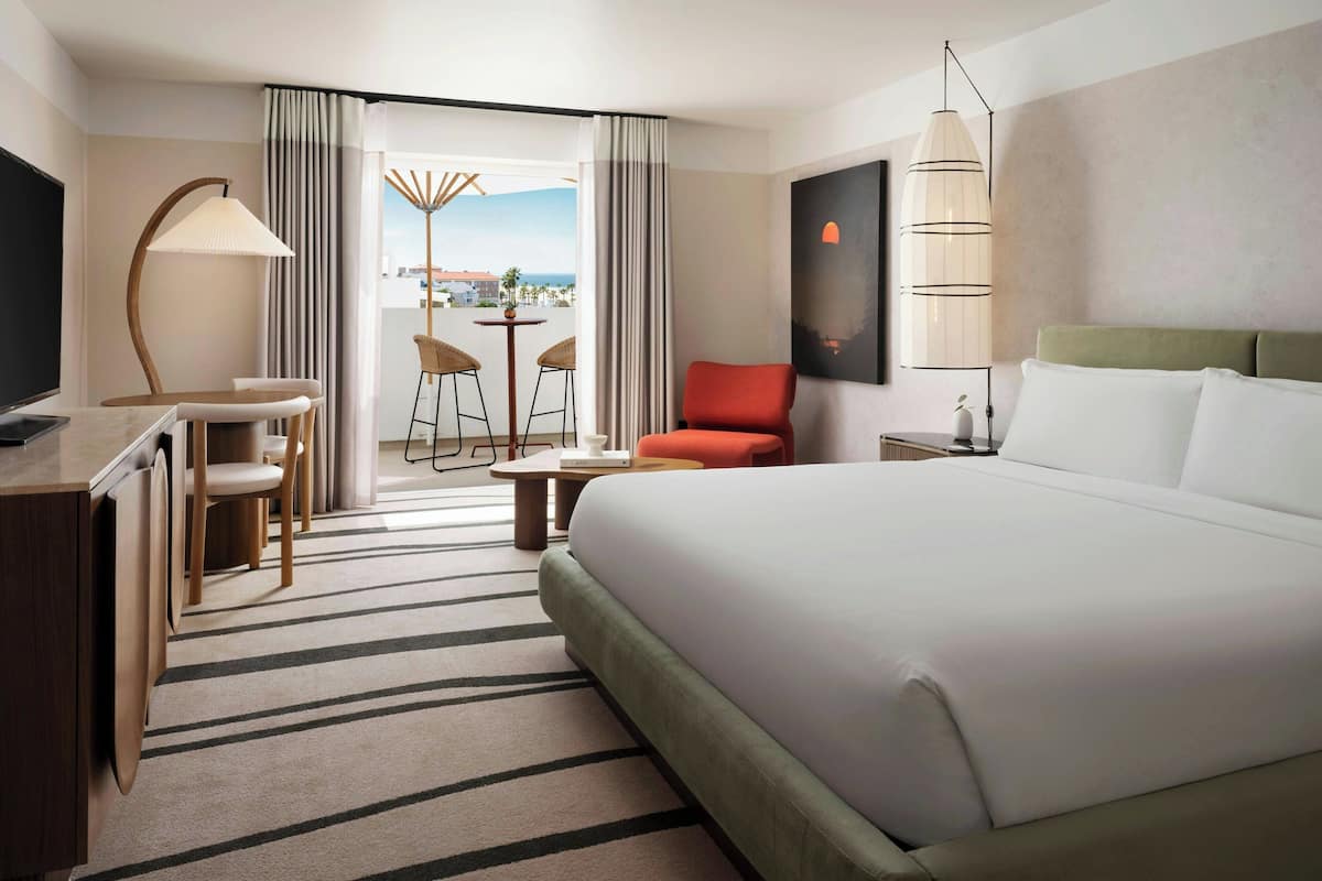 A hotel room with a large bed, a desk with a chair, a small table, and a view of the outside.
