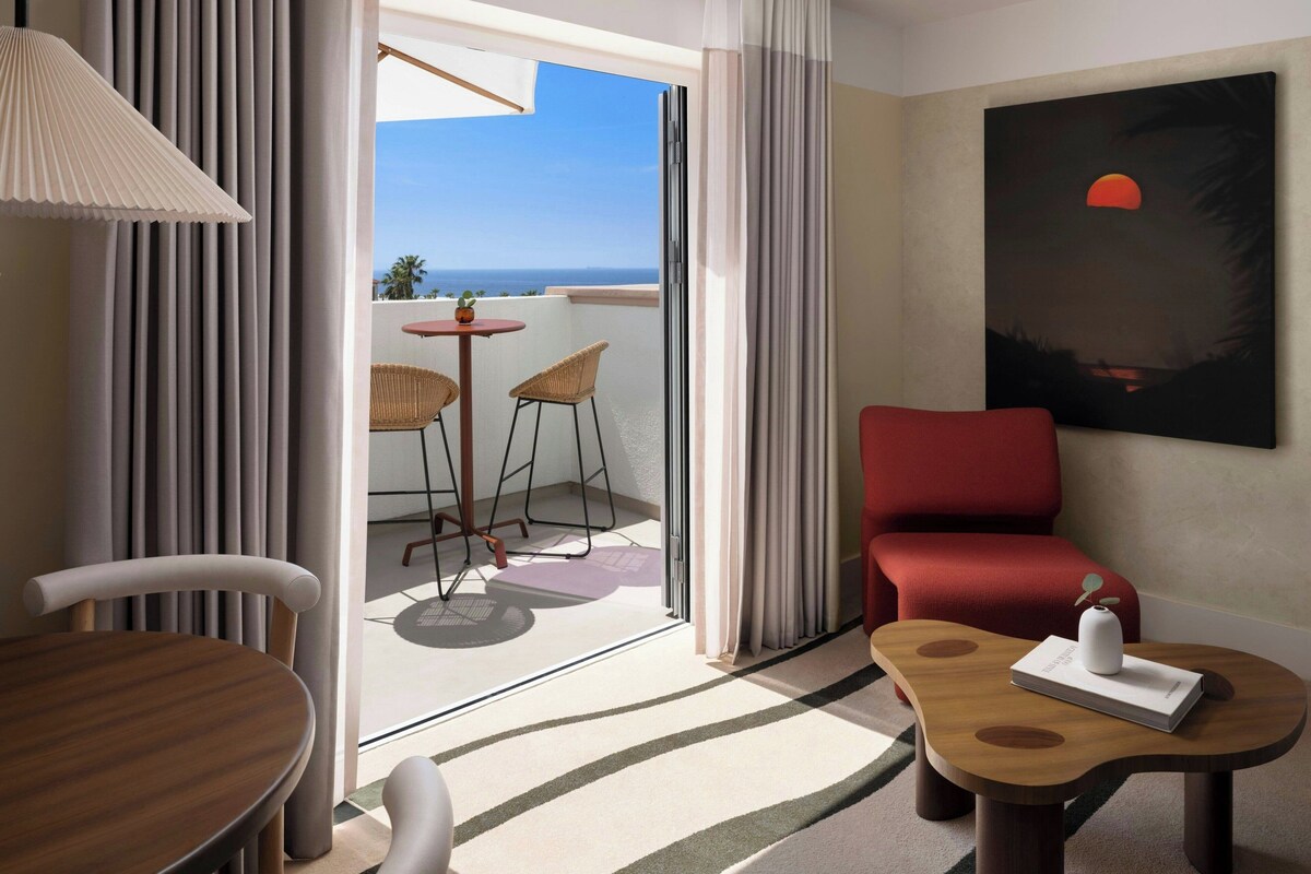 Habitación de hotel con balcón, una mesa de comedor, una silla roja, una pequeña mesa con un florero y un cuadro de una puesta de sol.