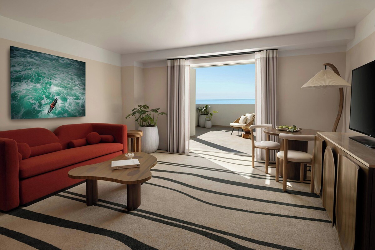 Habitación de hotel moderna con sofá rojo, una mesa de centro de madera, televisión, comedor con sillas, un ventanal con cortinas y una planta en maceta.