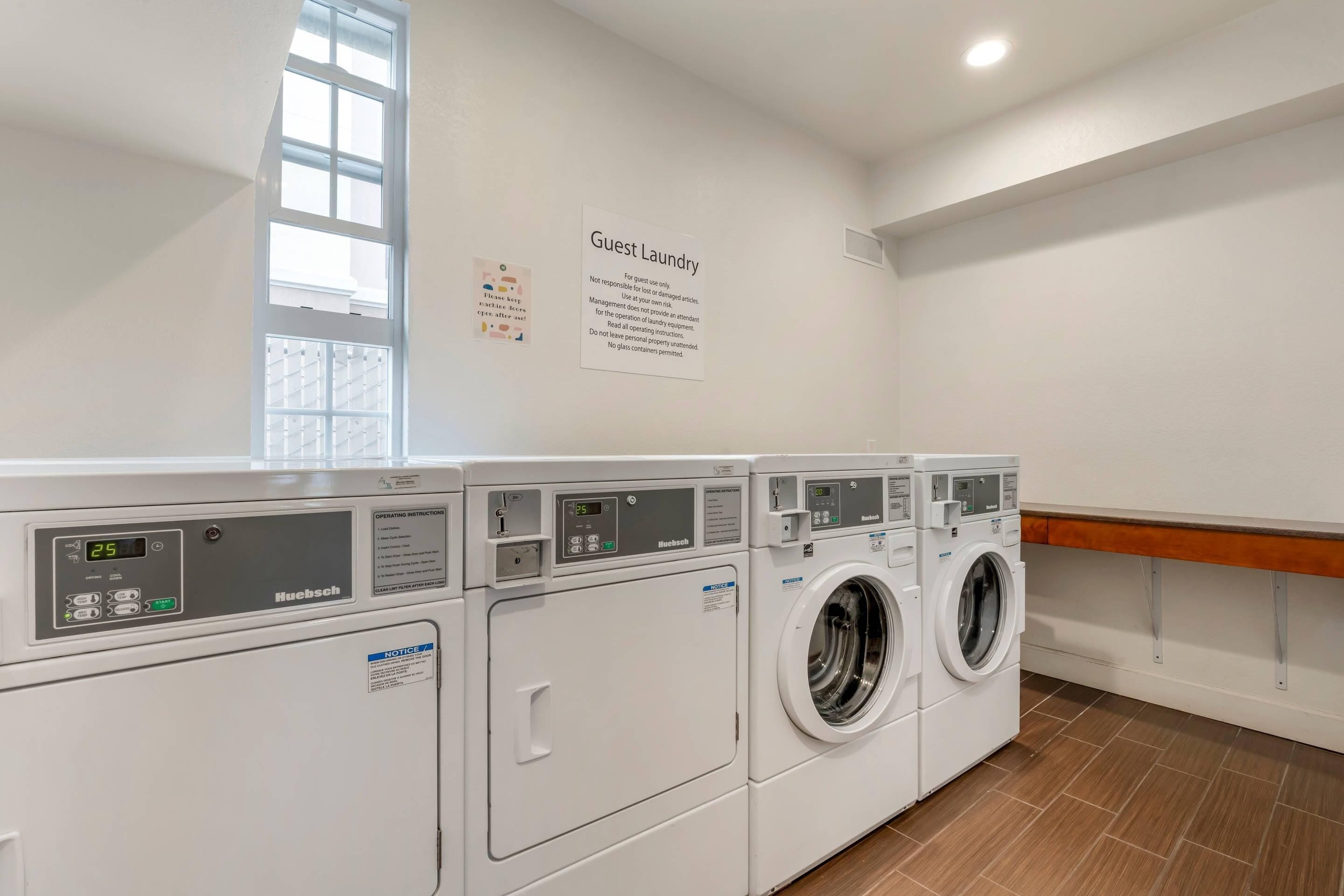A hotel laundry room with multiple washing machines and dryers, a sign indicating guest laundry use, and a window providing natural light.
